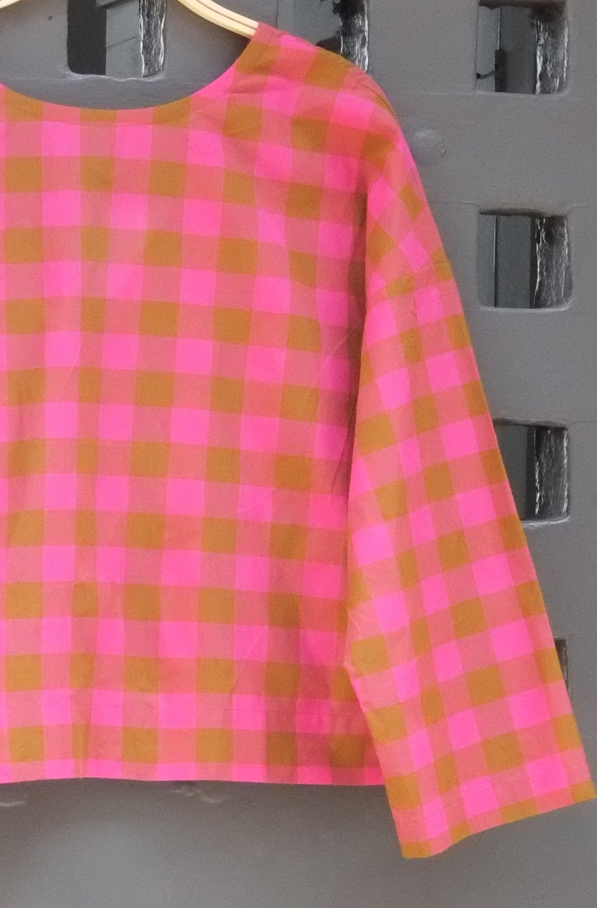 close up of pink and green checked long sleeve top pictures hanging on a wooden hanger on a grey door