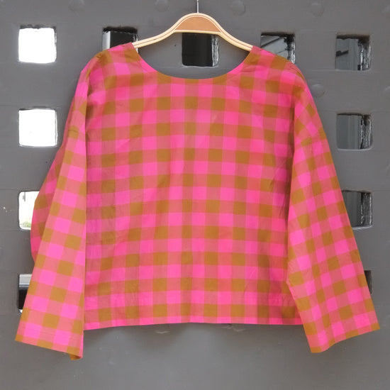 A pink and green gingham check long-sleeve top hanging on a wooden hanger against a grey lattice door