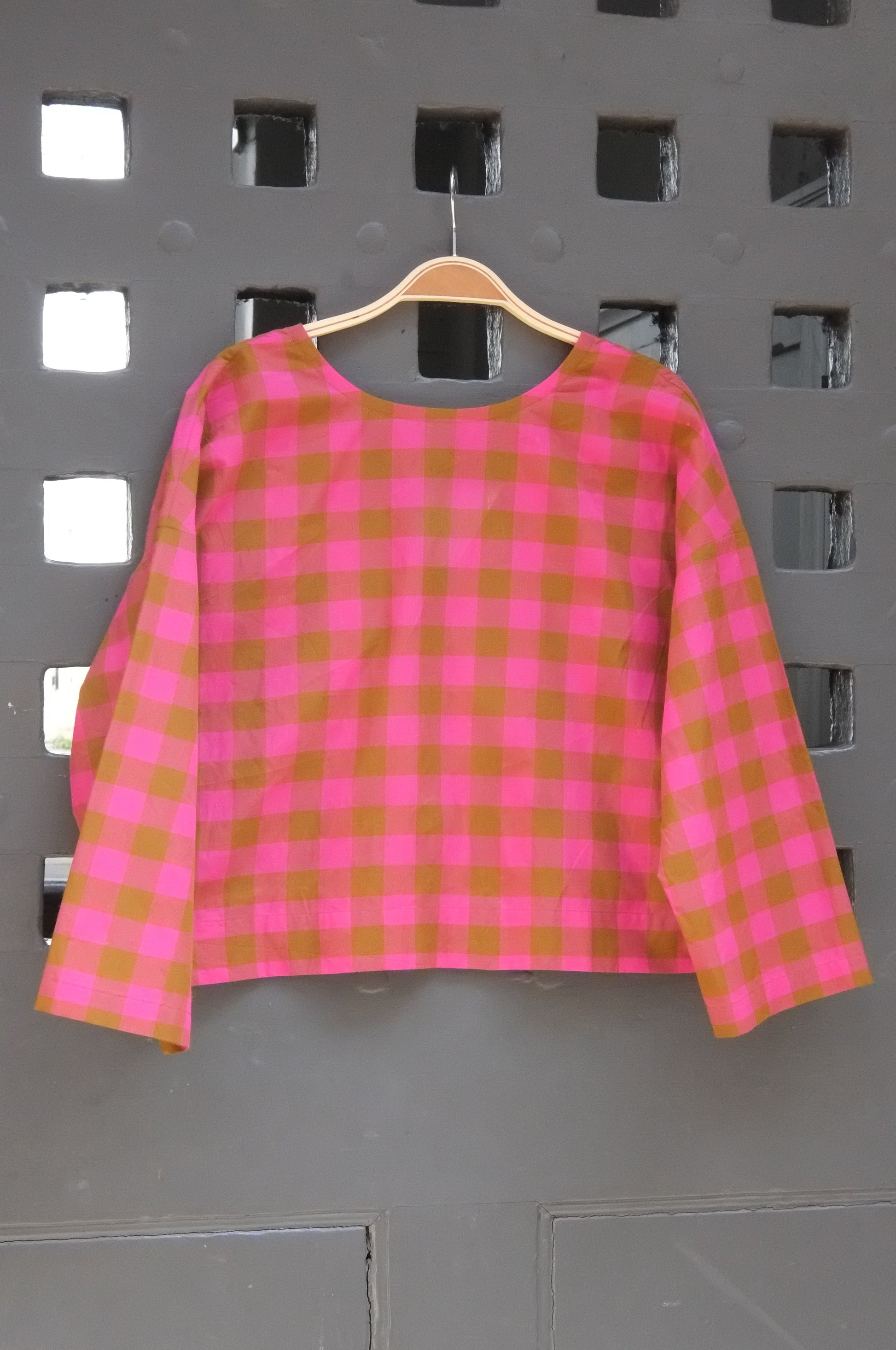 A pink and green gingham check long-sleeve top hanging on a wooden hanger against a grey lattice door