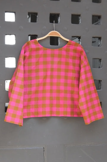 A pink and green gingham check long-sleeve top hanging on a wooden hanger against a grey lattice door