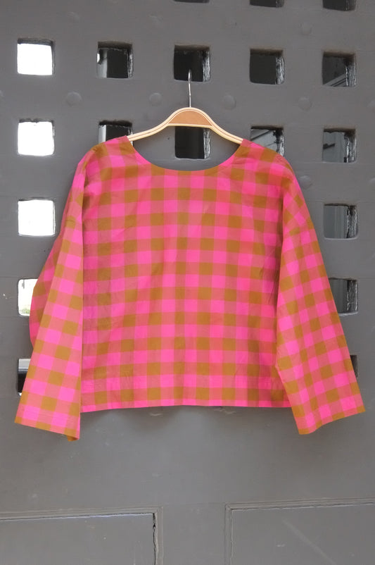 A pink and green gingham check long-sleeve top hanging on a wooden hanger against a grey lattice door
