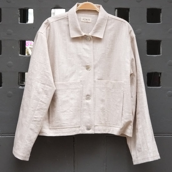 natural coloured workwear jacket with buttons photographed hanging on a grey door 