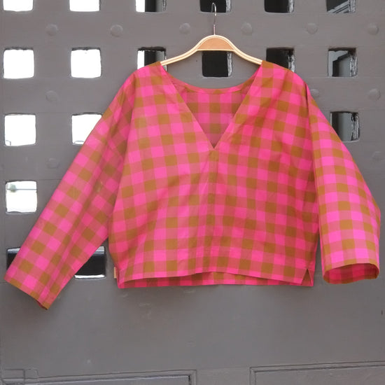 A pink and green gingham long-sleeve top hanging on a wooden hanger against a grey grid door