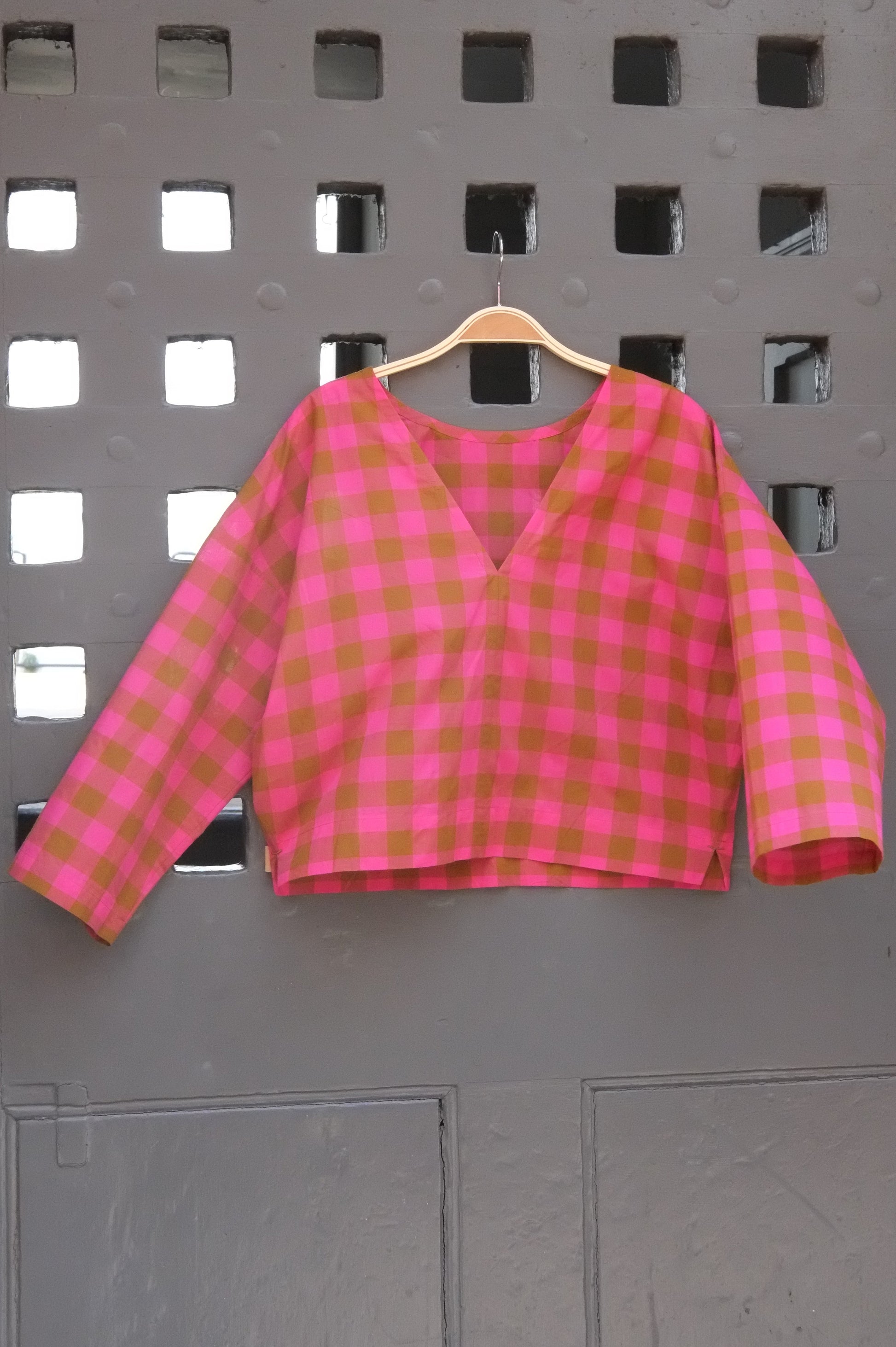 A pink and green gingham long-sleeve top hanging on a wooden hanger against a grey grid door