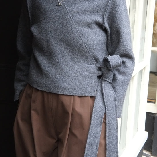 A woman wearing a grey wrap-style knit sweater with a side tie detail and brown wide-leg trousers, standing in front of a shop window.