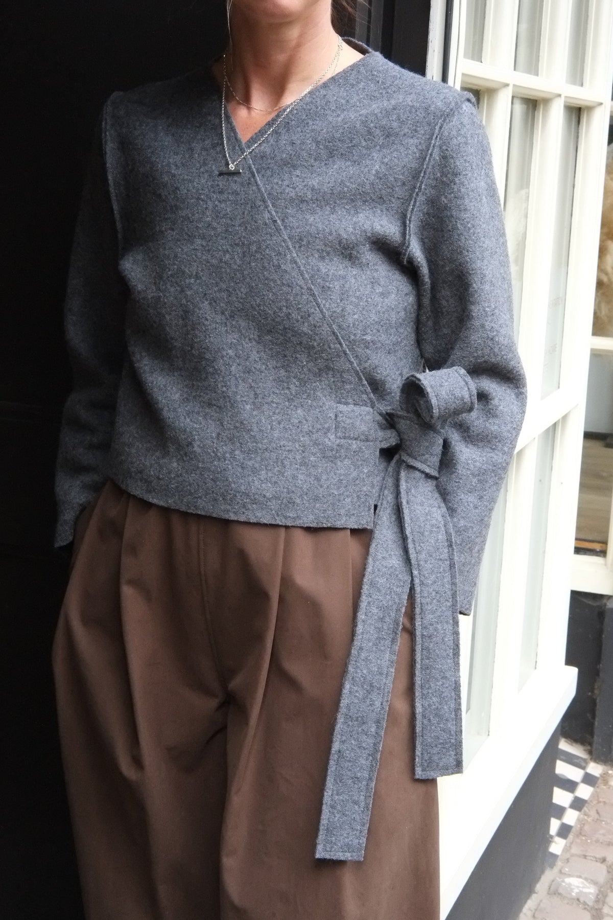 A woman wearing a grey wrap-style knit sweater with a side tie detail and brown wide-leg trousers, standing in front of a shop window.