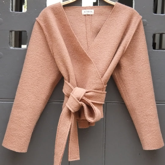 A camel wrap cardigan with long sleeves and a matching tie belt, hanging on a wooden hanger against a dark gray metal door.