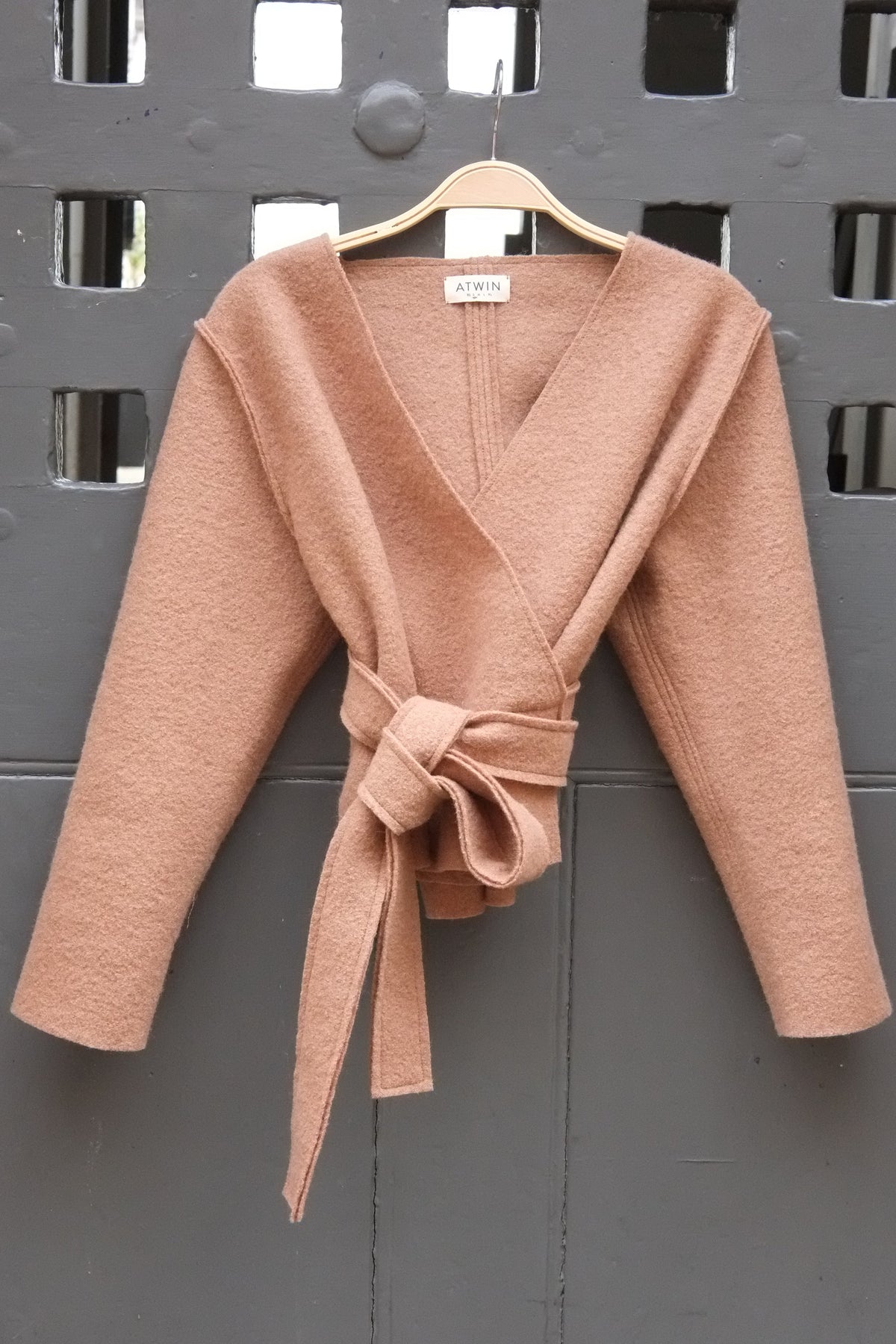 A camel wrap cardigan with long sleeves and a matching tie belt, hanging on a wooden hanger against a dark gray metal door.