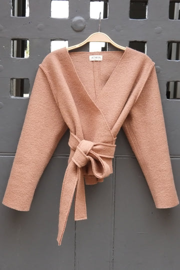 A camel wrap cardigan with long sleeves and a matching tie belt, hanging on a wooden hanger against a dark gray metal door.