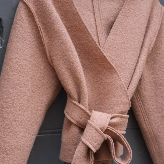 A camel wrap top with a tie belt, hanging on a wooden hanger against a dark grey paneled door. The brand label 'ATWIN' is visible at the collar.