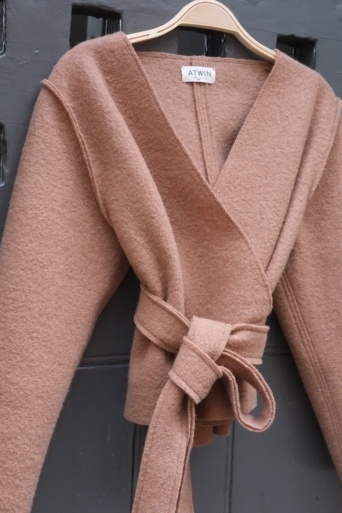 A camel wrap top with a tie belt, hanging on a wooden hanger against a dark grey paneled door. The brand label 'ATWIN' is visible at the collar.