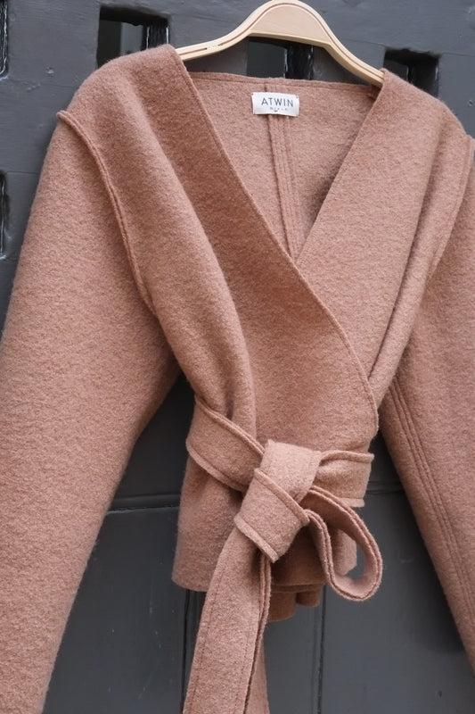 A camel wrap top with a tie belt, hanging on a wooden hanger against a dark grey paneled door. The brand label 'ATWIN' is visible at the collar.