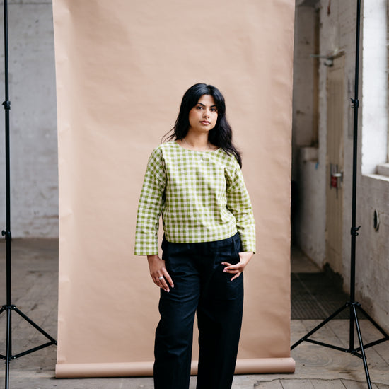 Woman in a green checkered shirt and black pants standing in front of a beige backdrop in an industrial setting.