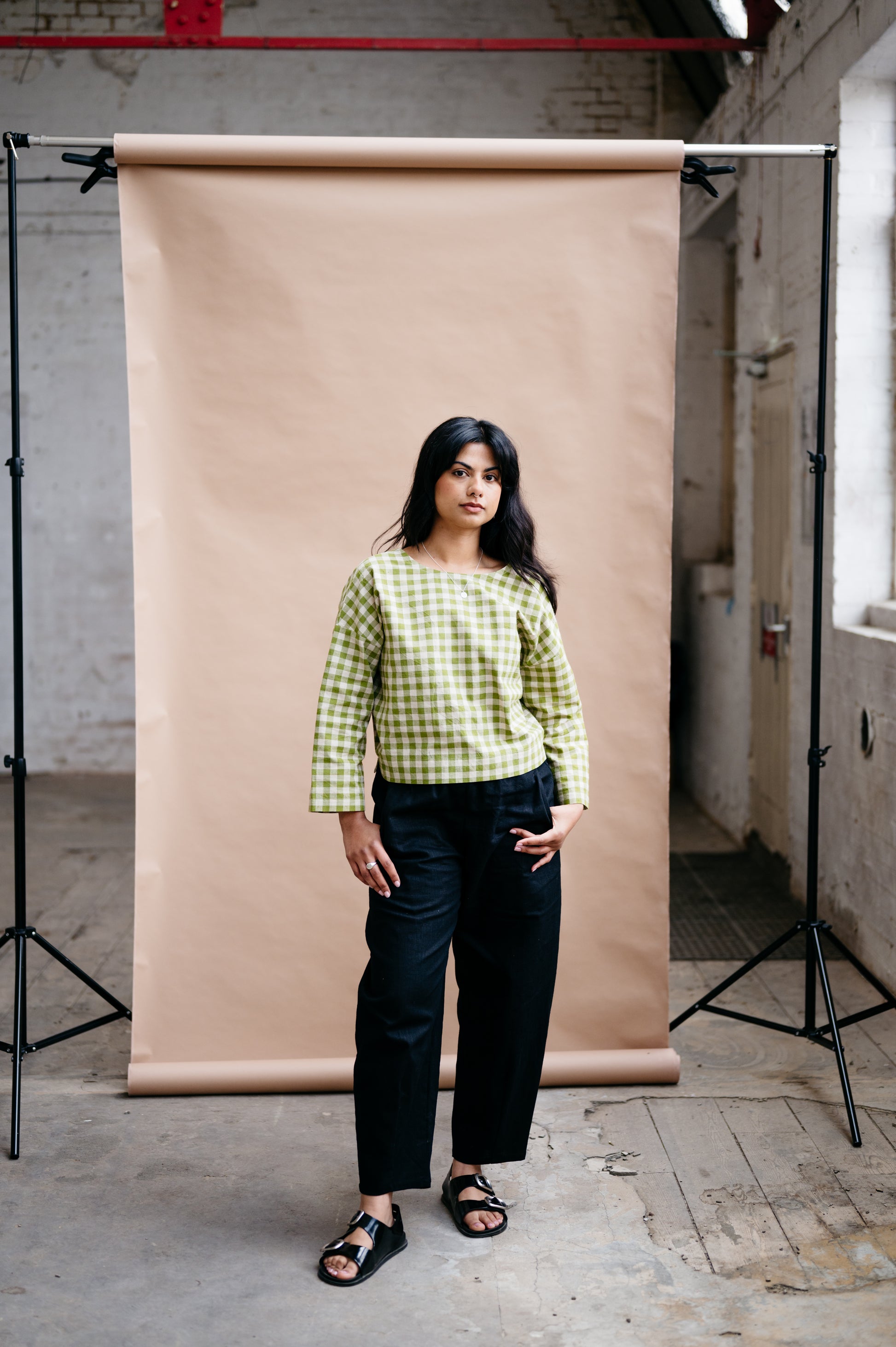 Woman in a green checkered shirt and black pants standing in front of a beige backdrop in an industrial setting.
