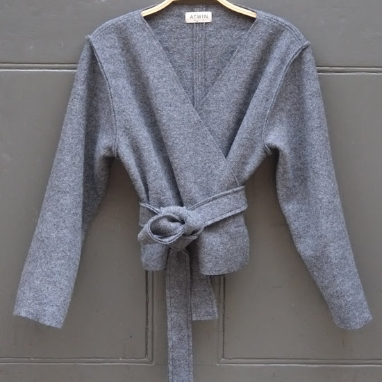 A grey wrap cardigan sweater with a tie waist, hanging on a wooden hanger against a dark gray metal door.