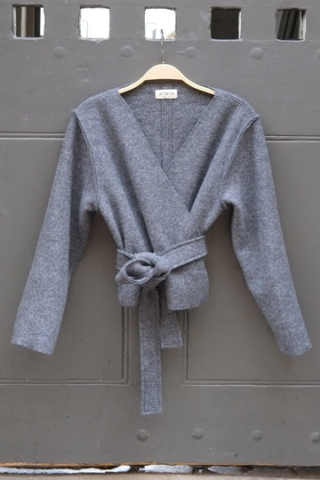 A grey wrap cardigan sweater with a tie waist, hanging on a wooden hanger against a dark gray metal door.