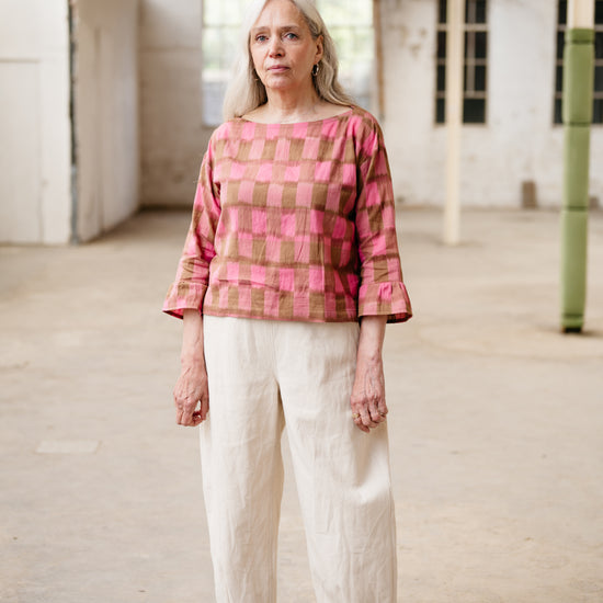 Woman wearing a pink and brown checkered top with white pants in an industrial setting.