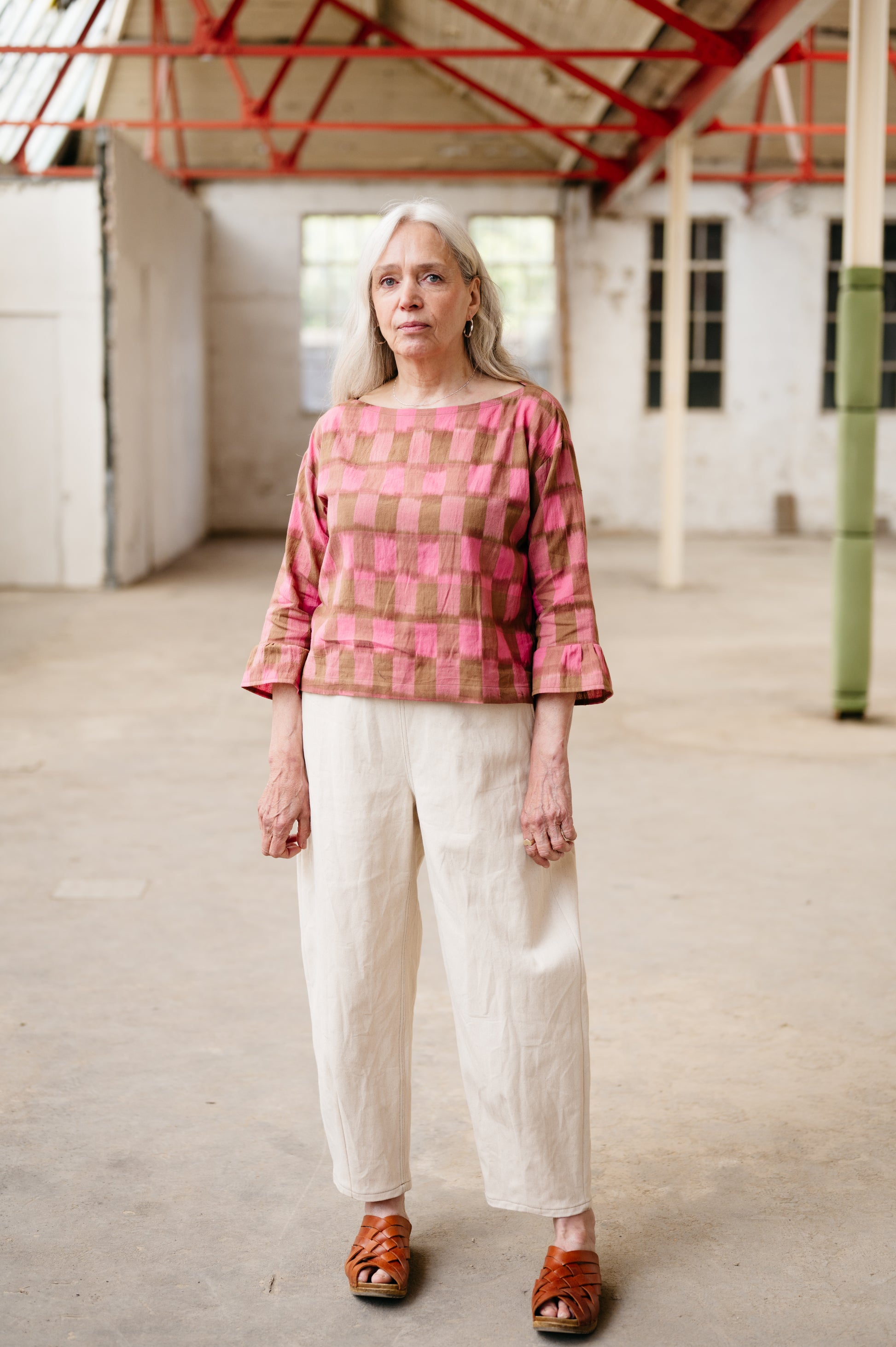 Woman wearing a pink and brown checkered top with white pants in an industrial setting.
