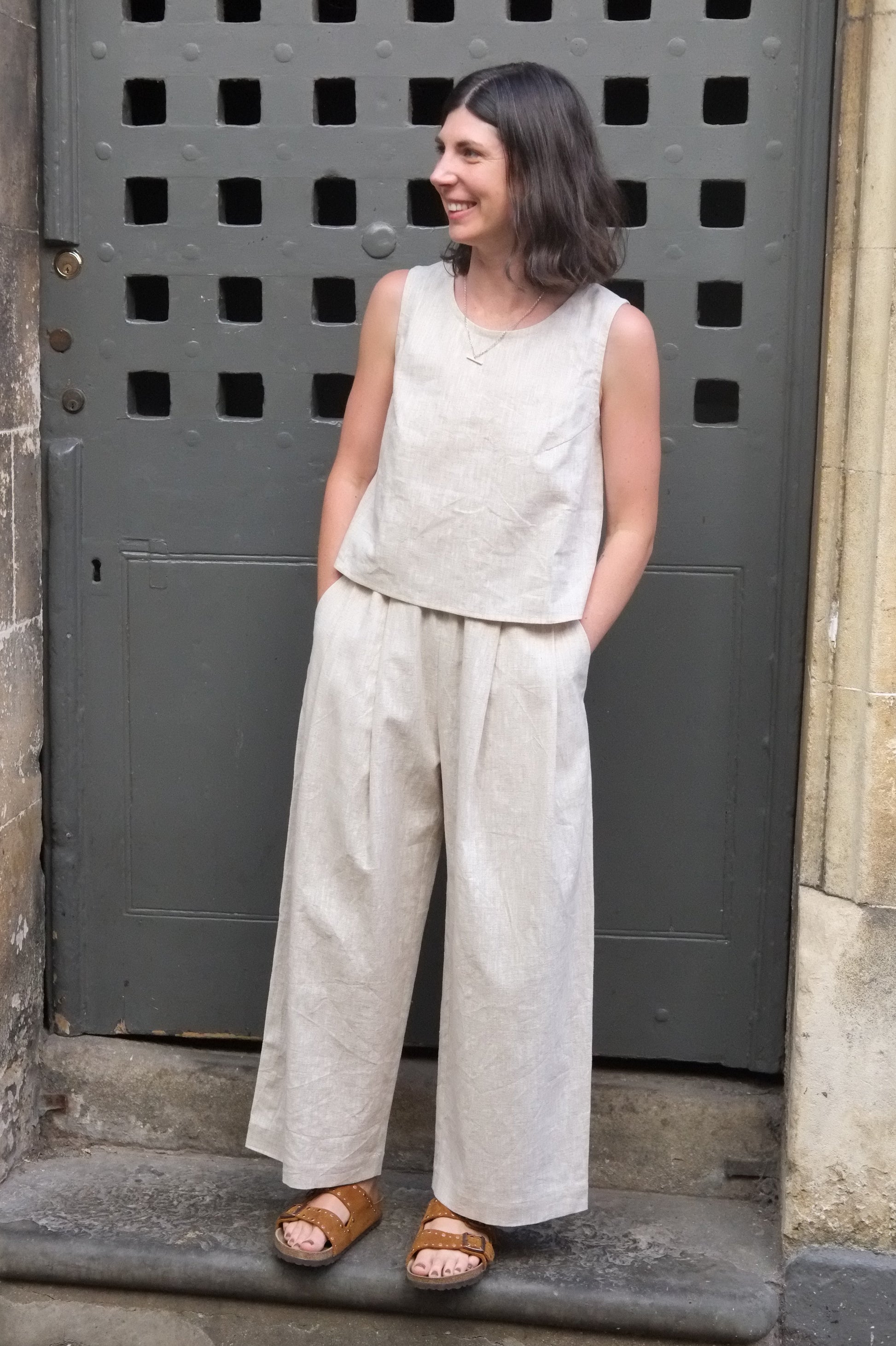 woman wearing a loose fitting cream coloured outfit with hands in pockets standing in front of an old door