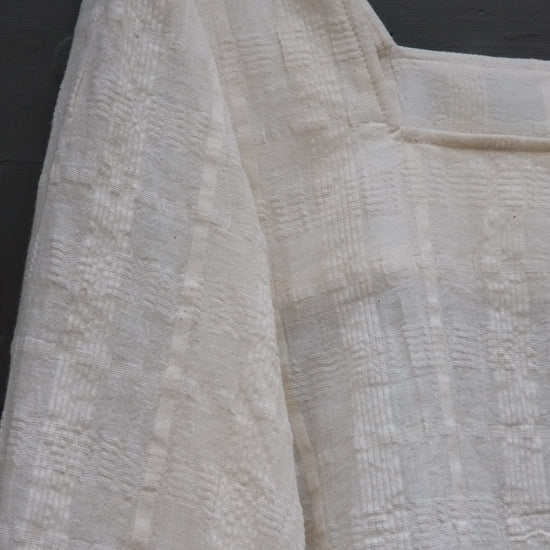 A close-up of a textured off-white or cream fabric garment, showing the woven pattern and folds.