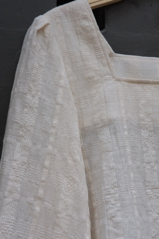 A close-up of a textured off-white or cream fabric garment, showing the woven pattern and folds.