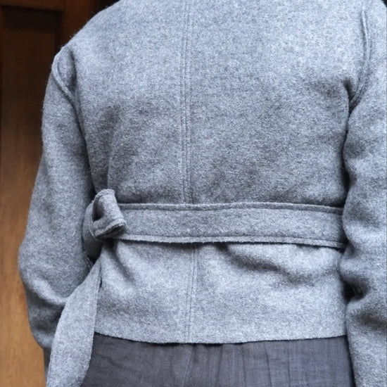 A person shown from the back wearing a light gray belted jacket and dark gray trousers, standing in front of a wooden door.