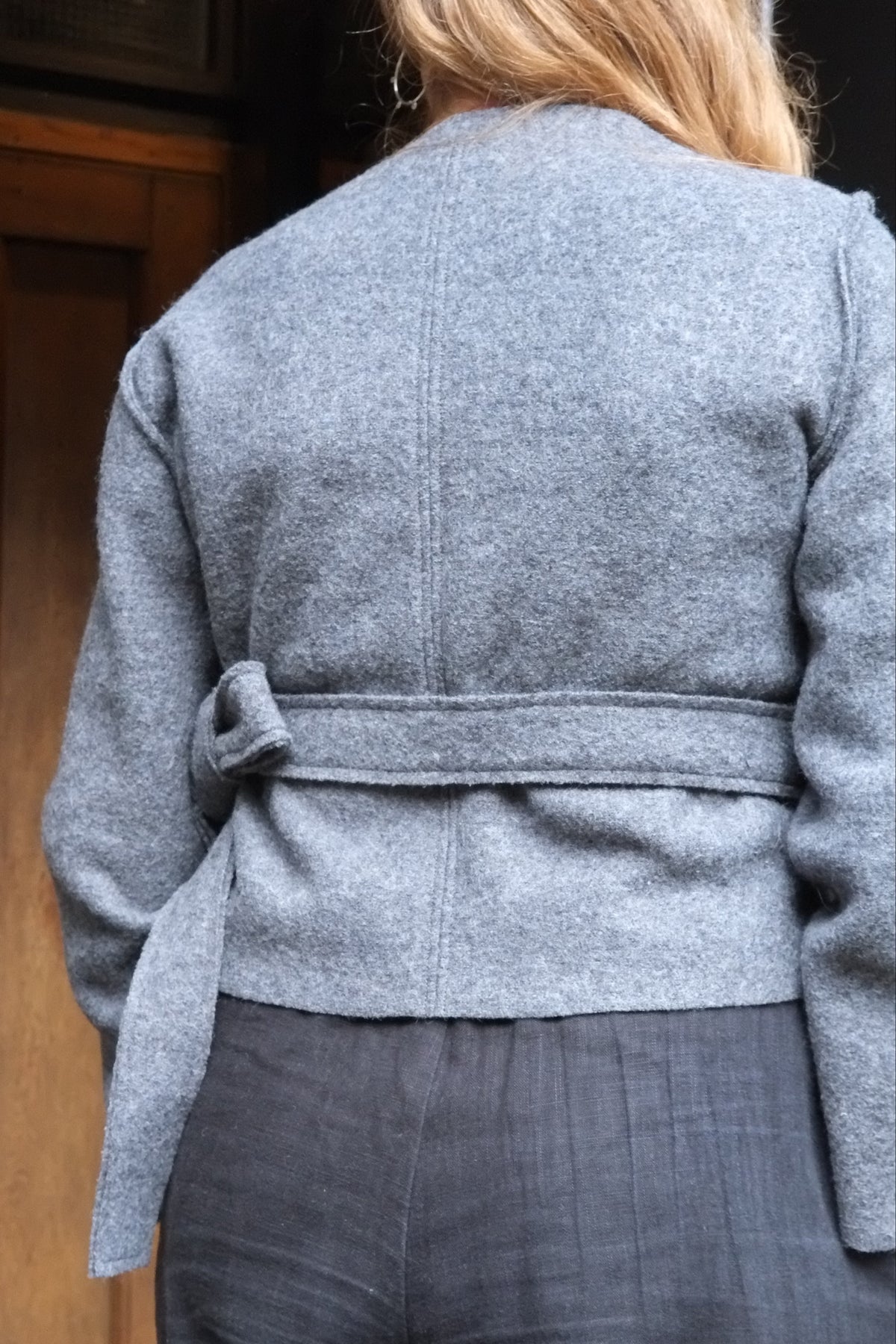 A person shown from the back wearing a light gray belted jacket and dark gray trousers, standing in front of a wooden door.
