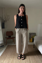 Load image into Gallery viewer, Woman wearing a black sleeveless top with white buttons and checkered pants in a living room.
