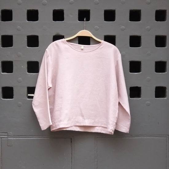 A light pink long-sleeve crewneck top hanging on a wooden hanger against a dark grey door with square cutouts.