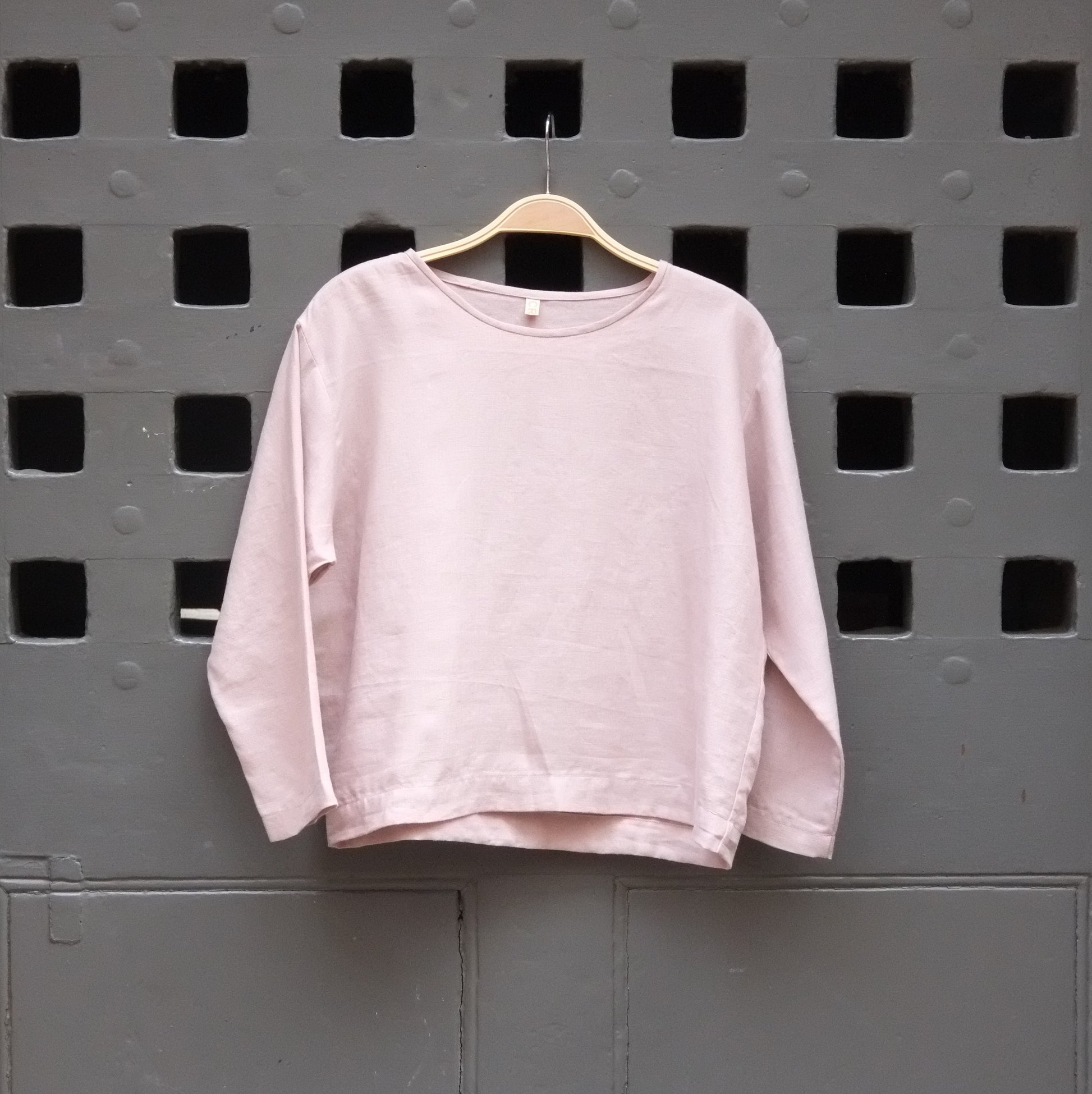 A light pink long-sleeve crewneck top hanging on a wooden hanger against a dark grey door with square cutouts.