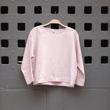 A light pink long-sleeve crewneck top hanging on a wooden hanger against a dark grey door with square cutouts.
