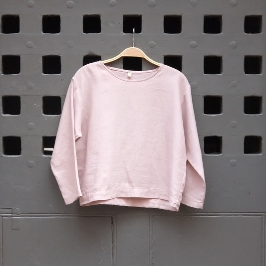 A light pink long-sleeve crewneck top hanging on a wooden hanger against a dark grey door with square cutouts.