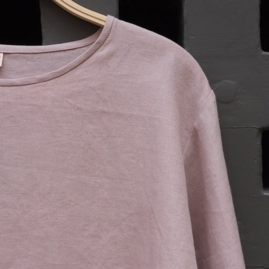 A dusty pink long-sleeve crewneck top hanging on a wooden hanger against a dark grey door.