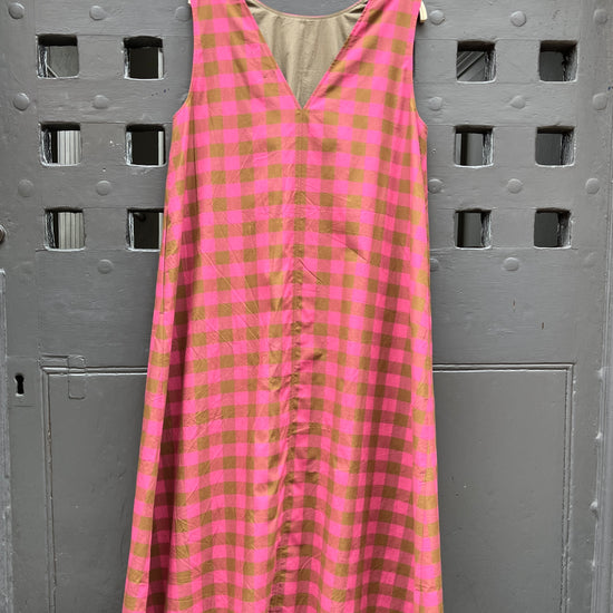 A sleeveless pink and olive green gingham check dress hanging on a wooden hanger against a gray door with square cutouts.