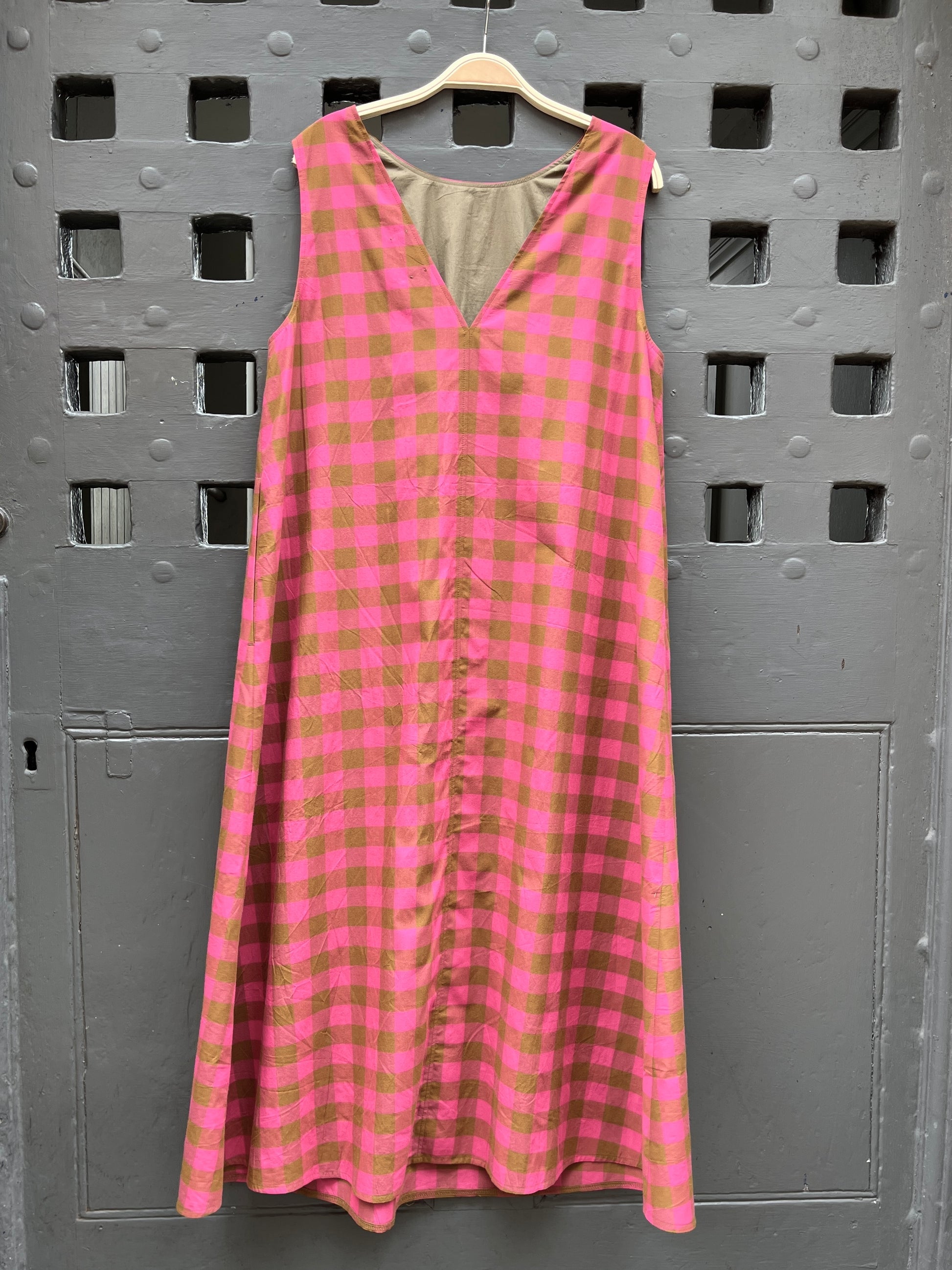 A sleeveless pink and olive green gingham check dress hanging on a wooden hanger against a gray door with square cutouts.