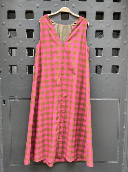 A sleeveless pink and olive green gingham check dress hanging on a wooden hanger against a gray door with square cutouts.