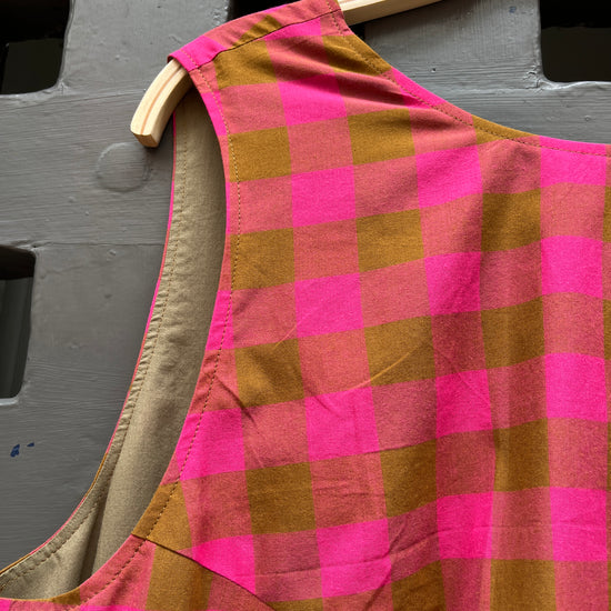 A close-up of a pink and olive plaid garment, hanging on a light wood hanger against a grey textured door