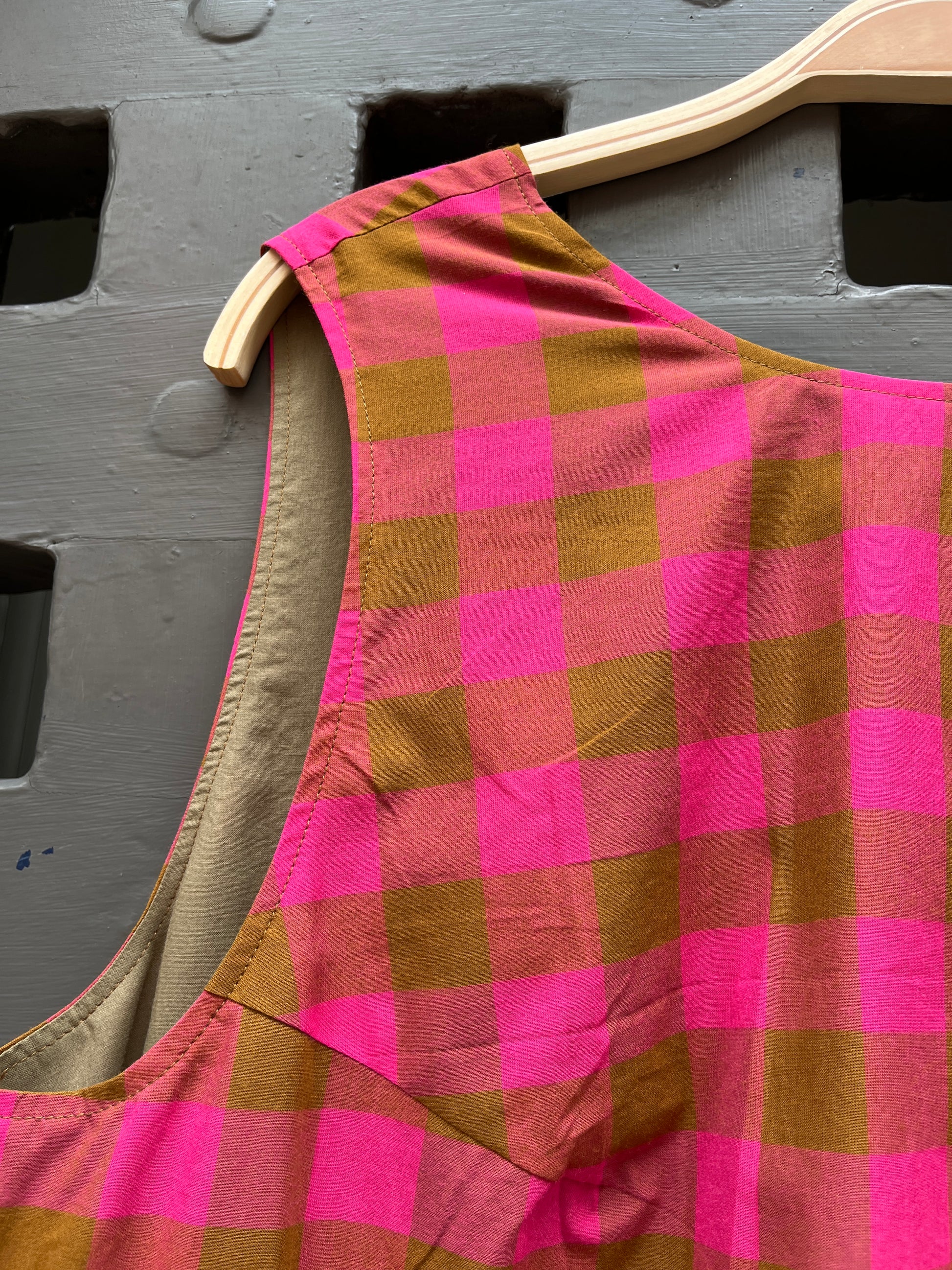 A close-up of a pink and olive plaid garment, hanging on a light wood hanger against a grey textured door