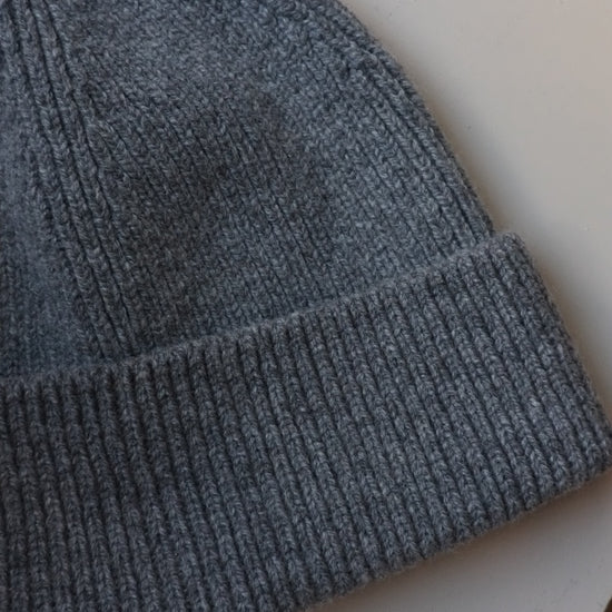 A close-up of a dark gray ribbed knit beanie hat, showing the texture of the yarn and the folded cuff, resting on a light off-white surface.