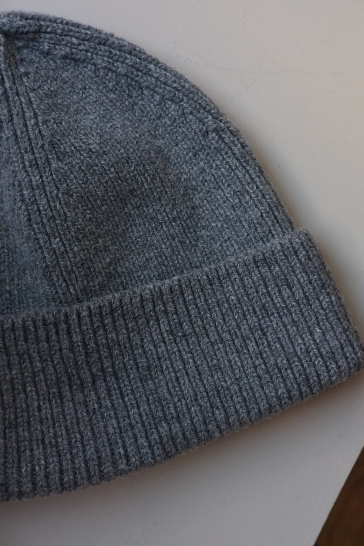 A close-up of a dark gray ribbed knit beanie hat, showing the texture of the yarn and the folded cuff, resting on a light off-white surface.