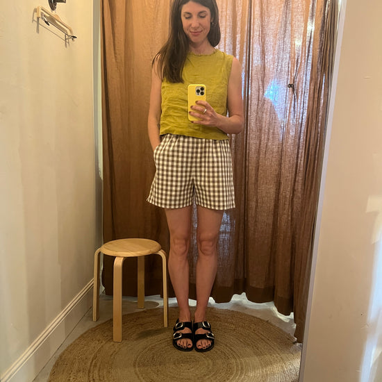 Woman taking a mirror selfie wearing a yellow top and checkered skirt in a room with wooden flooring.