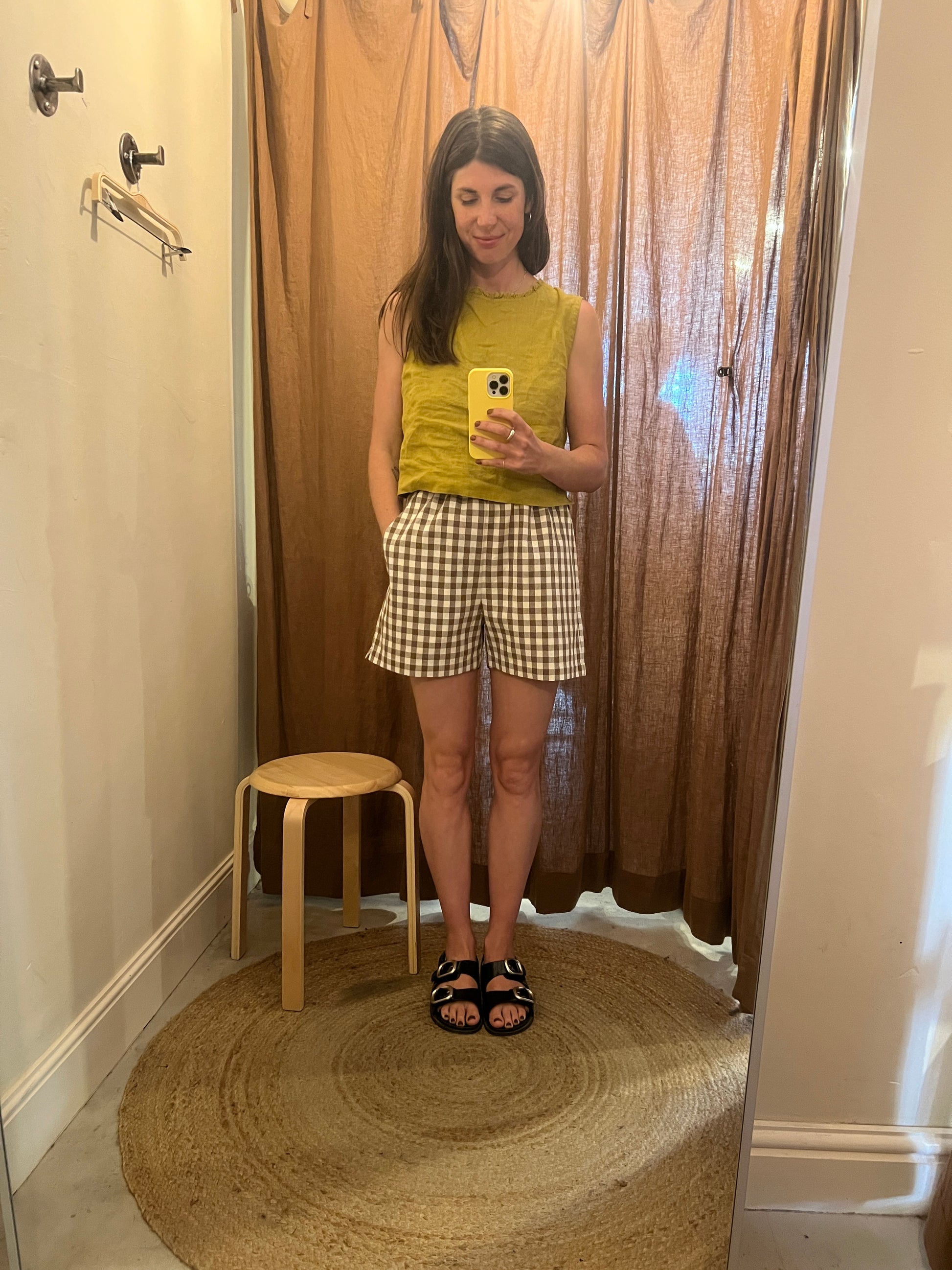 Woman taking a mirror selfie wearing a yellow top and checkered skirt in a room with wooden flooring.