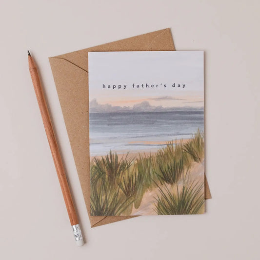 Greeting card with beach scene and 'happy father's day' text, accompanied by a pencil and brown envelope on a beige background.