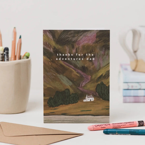 Greeting card with landscape illustration and 'thanks for the adventures dad' text, placed on a desk with stationery items.