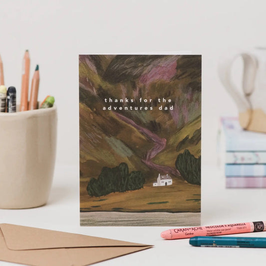 Greeting card with landscape illustration and 'thanks for the adventures dad' text, placed on a desk with stationery items.