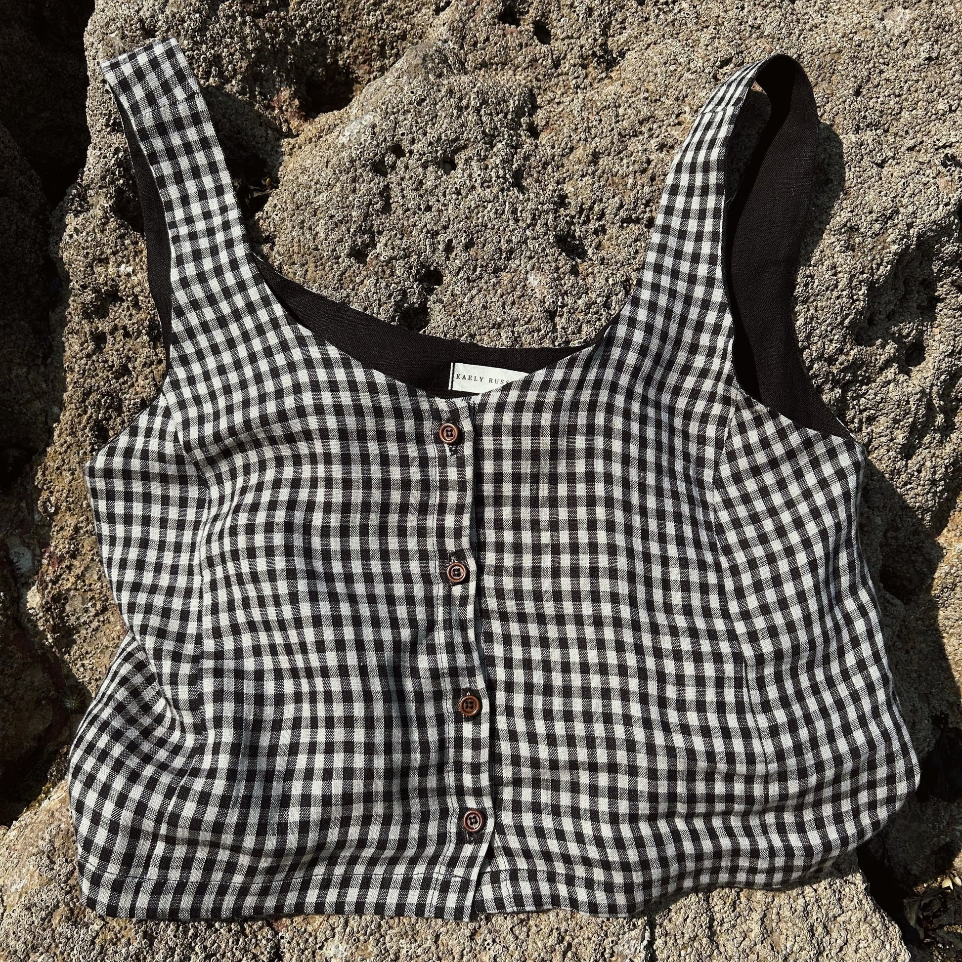A black and white gingham cropped tank top with brown buttons, laid flat on a textured gray stone surface.