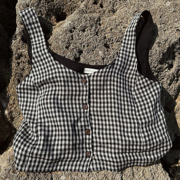 A black and white gingham cropped tank top with brown buttons, laid flat on a textured gray stone surface.