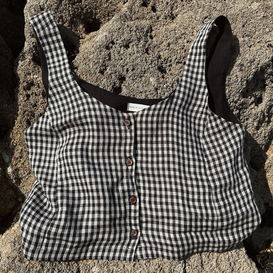A black and white gingham cropped tank top with brown buttons, laid flat on a textured gray stone surface.