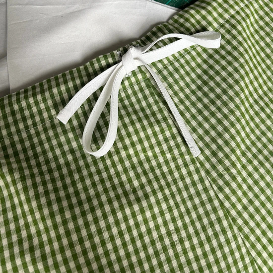 A close-up of a green and white gingham fabric item tied with a white ribbon, placed on a green cutting mat with a white fabric piece underneath.
