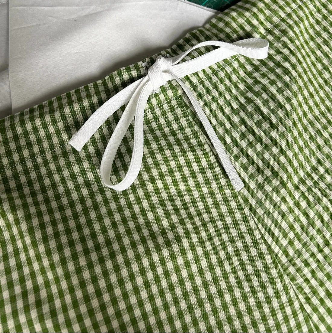 A close-up of a green and white gingham fabric item tied with a white ribbon, placed on a green cutting mat with a white fabric piece underneath.